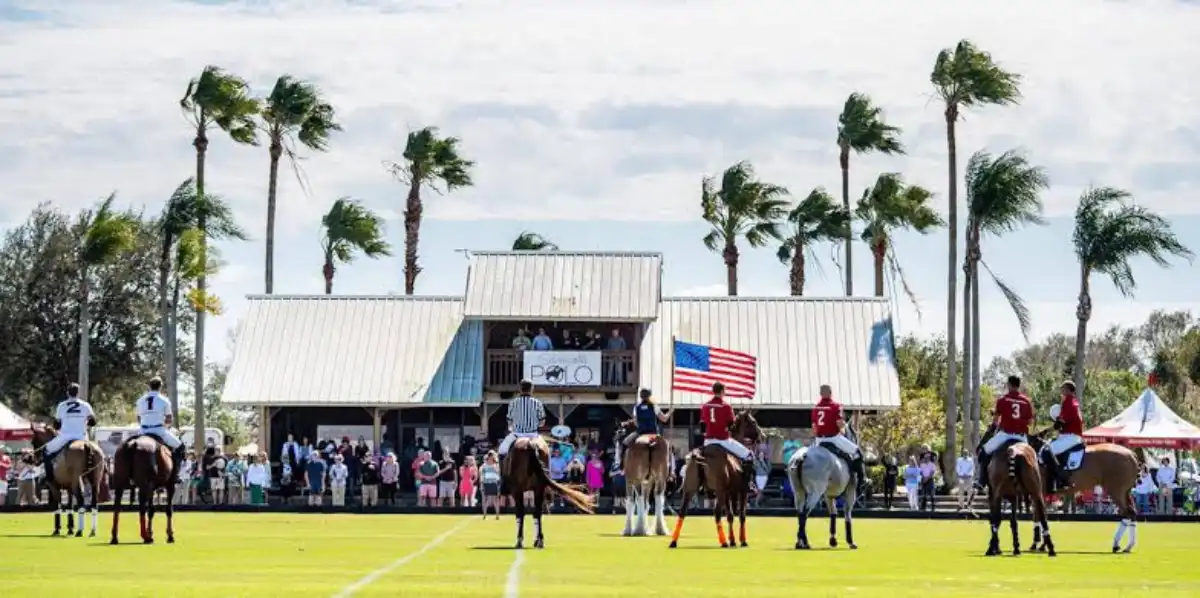 Polo players competing on a sunny field during a polo match in Myakka City, Florida, with spectators watching from the clubhouse and palm trees in the background.