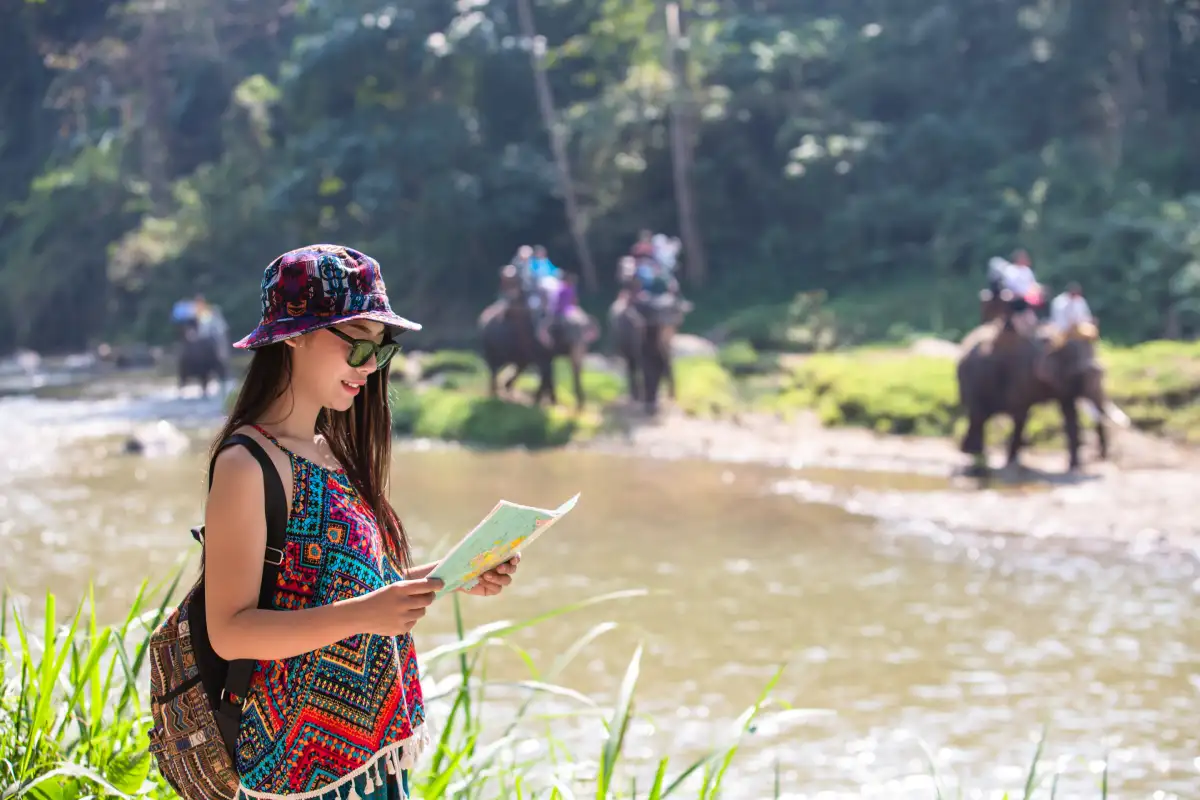 A woman in colorful traditional attire standing beside a river while using her phone, with elephants carrying riders crossing the water in the background surrounded by lush greenery.